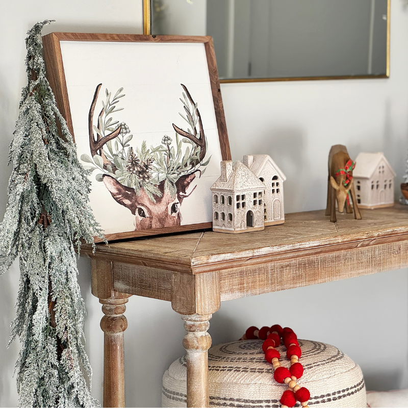 Holiday entryway table ideas: a close up shot of our Christmas console table featuring a frosted faux tree, small christmas village houses, and a wooden framed print of a reindeer! Cute and easy holiday decor for the season.