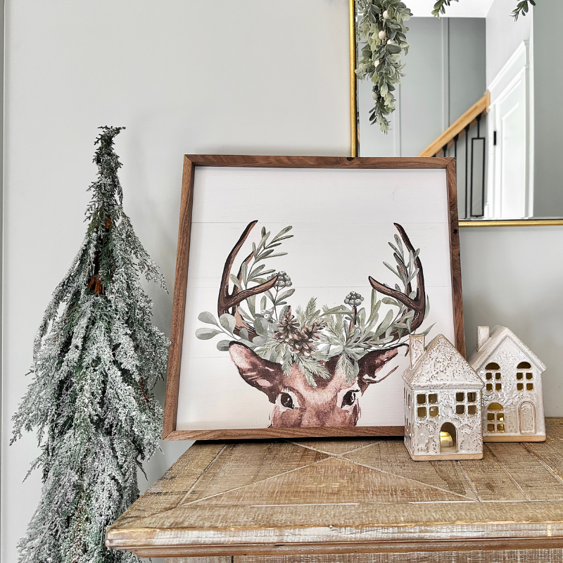 Holiday entryway table ideas: a close up shot of our Christmas console table featuring a frosted faux tree, small christmas village houses, and a wooden framed print of a reindeer! Cute and easy holiday decor for the season.
