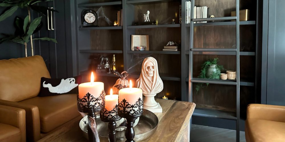 A cozy corner of a dark academia-inspired library, designed by Kadilak Homes, featuring Benjamin Moore Cheating Heart walls. The centerpiece of the coffee table includes gothic candle holders with flickering candles, a small bat-topped pumpkin, and a skeletal bust. In the background, a leather armchair with a ghost pillow sits beside dark wooden bookshelves filled with vintage books, cobwebs, and spooky decor, illuminated by warm, candlelight for a hauntingly elegant vibe.