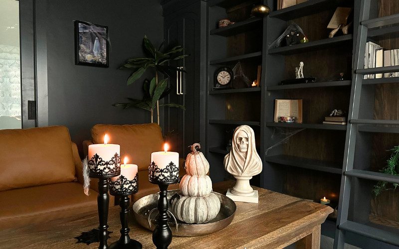 A view through a doorway into a haunted home library with dark academia decor, featuring a coffee table with pumpkins, skeletons, and candles, surrounded by dark wood shelves filled with spooky Halloween-themed accents.