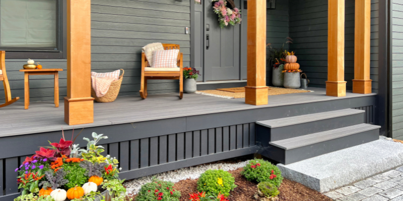 fall front porch ideas, dark house with black trim, porch with wood posts, porch with wood columns, hardie plank siding, house with black windows, front porch idea, cozy outdoor seating, pumpkin decor, front door with wreath, fall wreath ideas, set of planters, sitting area, west elm planter, wood outdoor rocking chairs, porch landscaping ideas with barrel, green artiscapes landscaping billerica ma