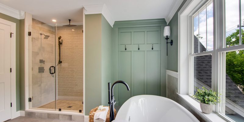 Primary bathroom with a soaking tub and black floor-mounted faucet, with a hanging towel hook against a sage green board and batten accent wall, and a large walk-in shower with four different shower heads, off white textured subway tile, and floor to ceiling glass doors