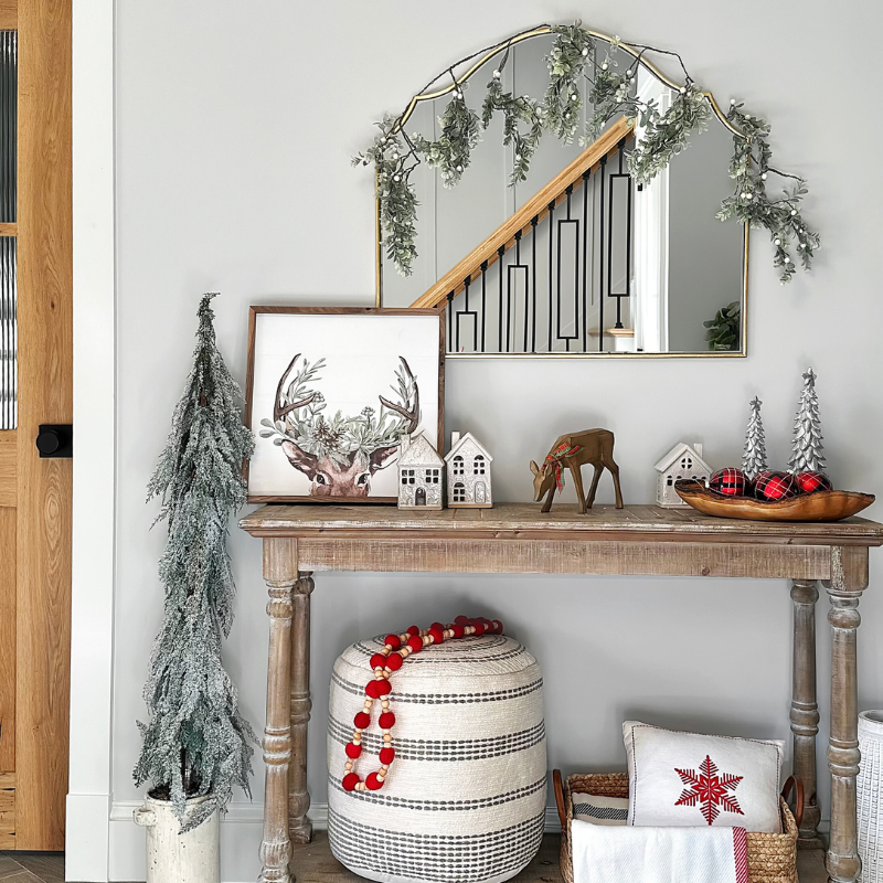 A photo of our entryway console table decorated for the holidays! We went with simple, neutral, and slightly modern Christmas decorations. A faux frosted tree, a pastel reindeer print, a gold framed mirror decorated with faux garland, ceramic christmas village houses, and a cozy holiday blanket and throw pillow.