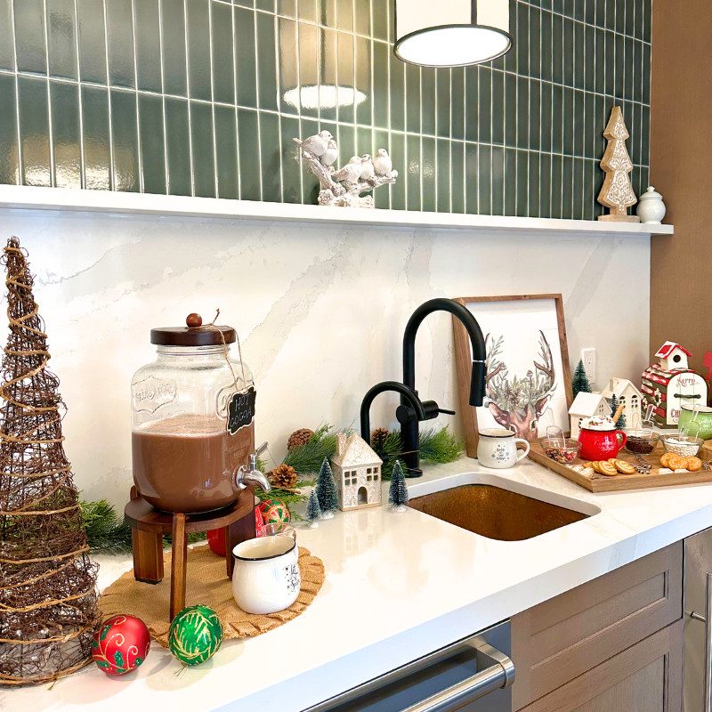 A festive hot chocolate bar setup on a white countertop with green backsplash tiles. The scene includes a hot chocolate dispenser on a wooden stand, holiday decorations like ornaments, a burlap mat, and ceramic houses, with a sink and black faucet in the background.