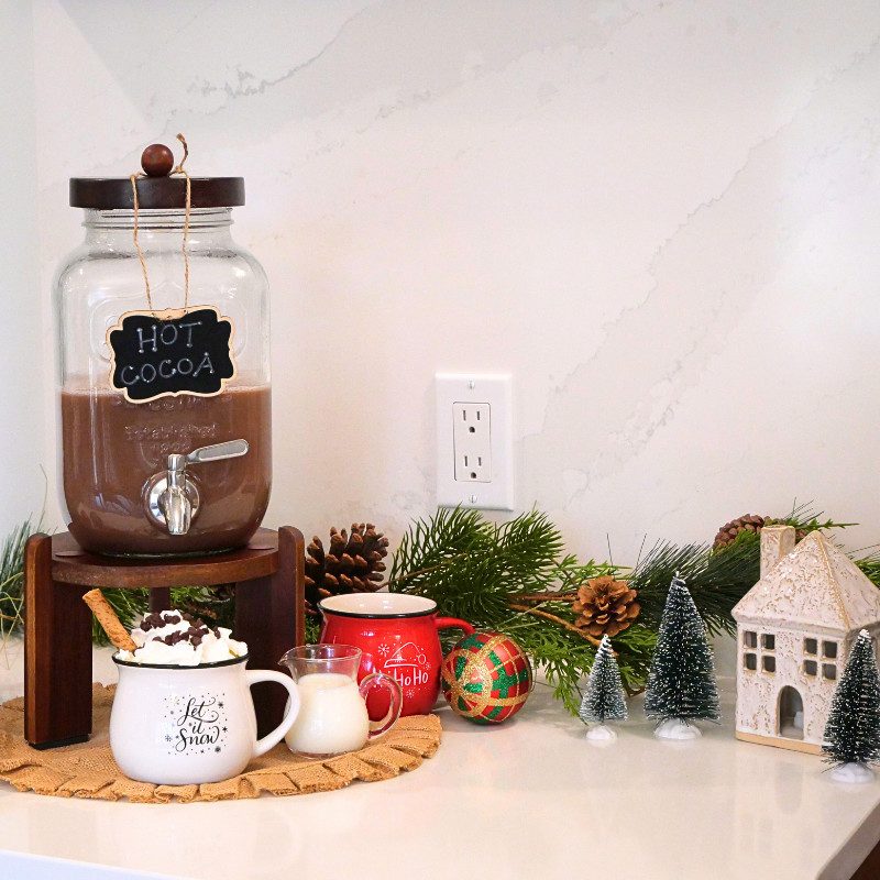 A close-up of a glass hot chocolate dispenser with a chalkboard label reading "Hot Cocoa," styled on a wooden stand with holiday accents like pinecones, greenery, and a cozy mug of whipped cream-topped hot chocolate.