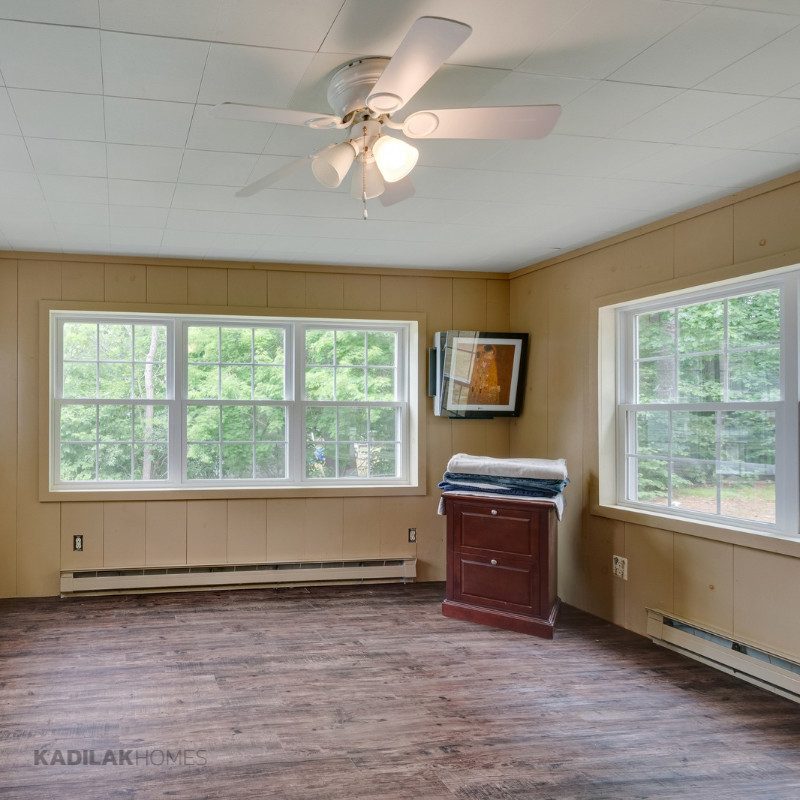Before family room renovation with wood panel walls, baseboard heaters, ceiling fan, and large windows in a Cape style home.