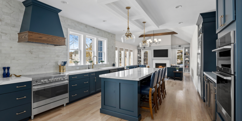 Navy blue kitchen cabinets with stained wood accents, neutral subway tile, and 2” thick quartz countertops. Kitchen with large island open to family room with chevron light box above. Navy blue kitchen cabinets. High end kitchen with induction range. Brushed gold kitchen pendant lighting with clear glass.