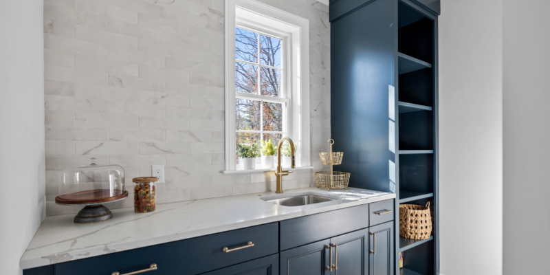 Navy blue kitchen cabinets with stained wood accents, neutral subway tile, and 2” thick quartz countertops. Kitchen with large island open to family room with chevron light box above. Navy blue kitchen cabinets. High end kitchen with induction range. Brushed gold kitchen faucet with marble subway tile backsplash inside patnry