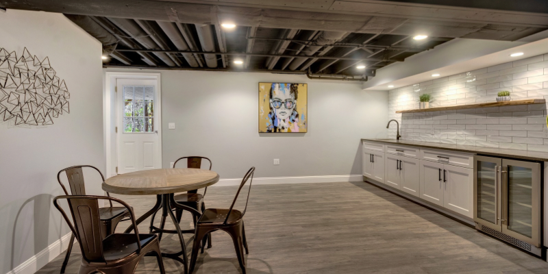 A spacious, modern basement area featuring an exposed black ceiling with beams and piping, complementing the dark, wood-look flooring. To the left, a simple dining area with a round table and four metal bistro chairs sits near a white door that leads outside, offering a touch of natural light. On the right, a sleek kitchenette boasts white cabinetry, stainless steel appliances including a wine fridge, and a long counter with a subway tile backsplash, illuminated by under-cabinet lighting. Above the kitchen area, recessed lights are strategically placed. A piece of abstract wire wall art adorns the wall to the left, while a colorful, contemporary painting of a figure with glasses adds a vibrant focal point on the adjacent wall. The room's neutral color palette is accented by these artistic elements, creating a cozy yet stylish space.