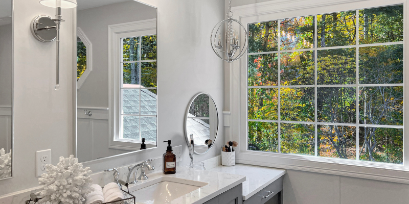 Long double vanity with sconces, double vanity with drop down make-up counter, vanity with chandelier, bathroom with chrome fixtures, large picture window in bathroom, benjamin moore bathroom walls Shoreline 1471, Sherwin Williams Serious Gray cabinets