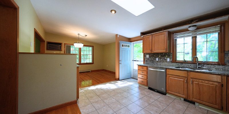 A dated kitchen featuring oak cabinets, beige tiled floors, and a granite countertop with a mosaic tile backsplash. The space includes a single window over the sink, allowing natural light to filter in. The room has a small entry door painted light blue, with outdated design elements such as a boxy wall partition and a low-hanging chandelier over the dining area. Yellow-toned walls and wood trim contribute to the old-fashioned feel, while the kitchen layout feels compact and closed off, awaiting a modern renovation.