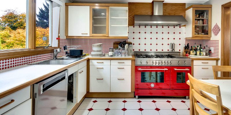 A mid-century modern kitchen with a vibrant red and white color scheme featuring a bold red range and white cabinetry with wooden trim. The backsplash and floor are adorned with matching red and white geometric tiles, adding a retro charm to the space. Large windows bring in natural light, enhancing the kitchen's bright and airy feel. The kitchen is a mix of functional elements and vintage style, showcasing a blend of practical design and nostalgic aesthetics.