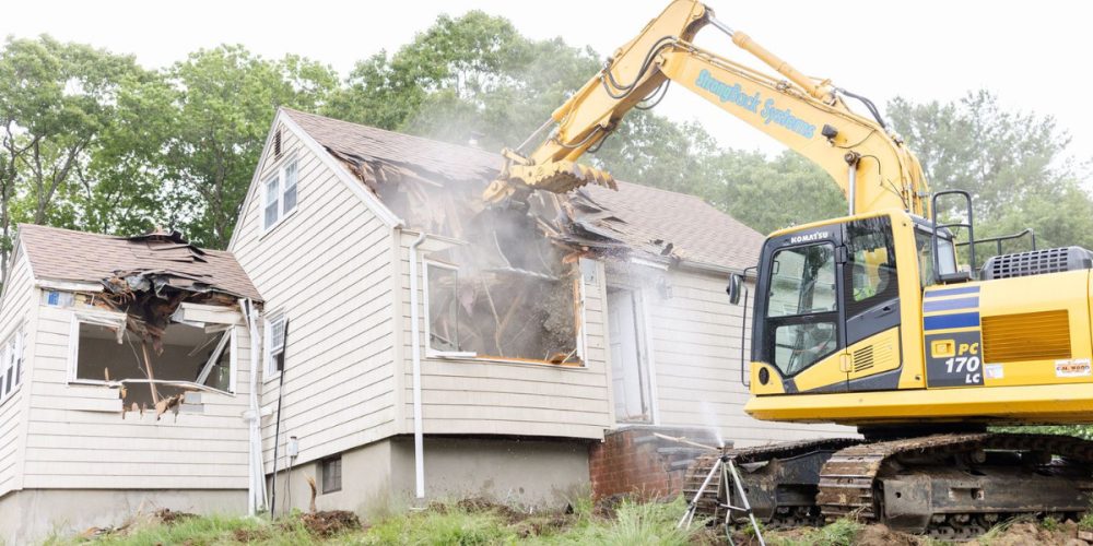 Yellow excavator demolishing a beige house with siding during the first phase of a home renovation project in Burlington, MA, with debris and dust in the air.