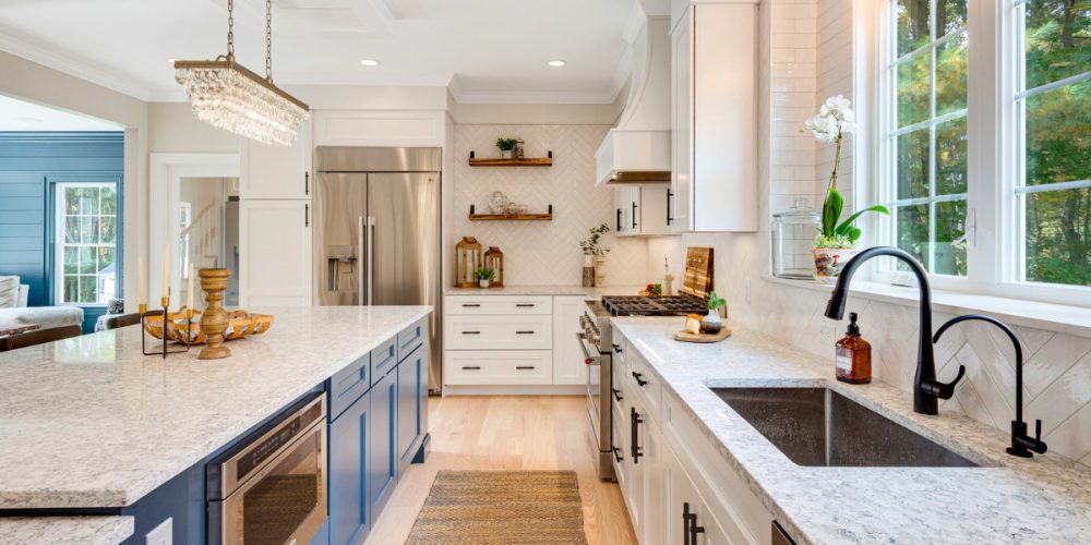 A spacious modern kitchen featuring blue lower cabinets and white upper cabinetry, adorned with white Calacatta quartz countertops. Elegant floating wood shelves, stainless steel appliances, and a crystal chandelier create a bright and inviting atmosphere.
