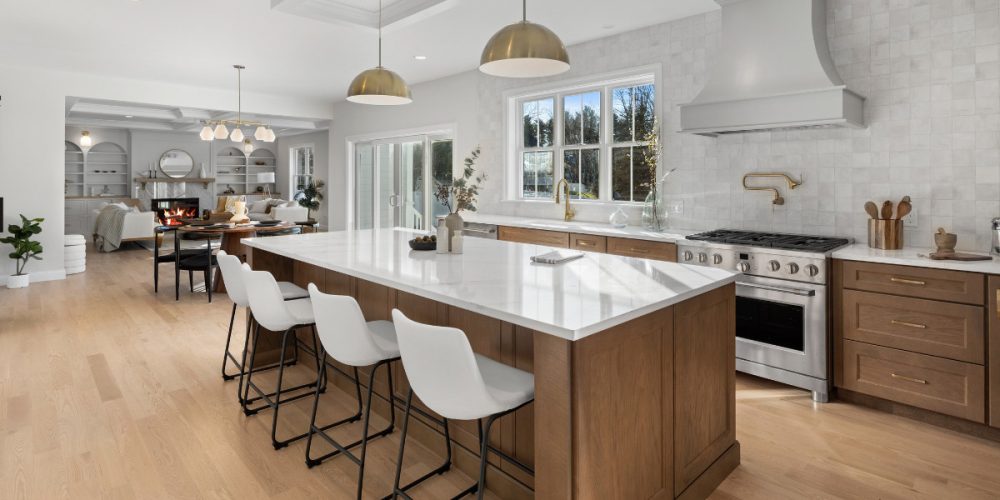 A modern kitchen featuring natural wood cabinets, white quartz countertops, and gold accents. The open floor plan connects the kitchen with a cozy living space, showcasing built-in shelving and a fireplace