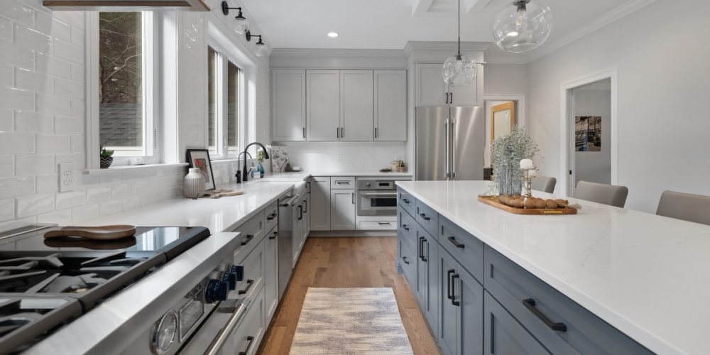 A sleek gray and blue kitchen design with a long kitchen island featuring white quartz countertops. The space is accented with white subway tile backsplash, stainless steel appliances, and large windows providing ample natural light.