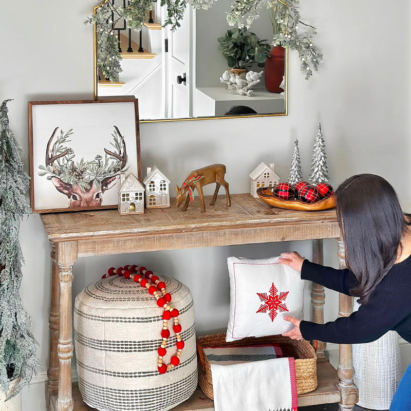 Susan Kadilak staging her entryway console table for the holidays! Featuring a reindeer art print, ceramic Christmas houses, a live edge olive wood bowl, a wool blend throw, a snowflake throw pillow, vintage garland, vintage ornaments, a braided basket, textured pouf, resin Christmas trees, terracotta vase, artificial HGTV tree, and white berry garland from Target!