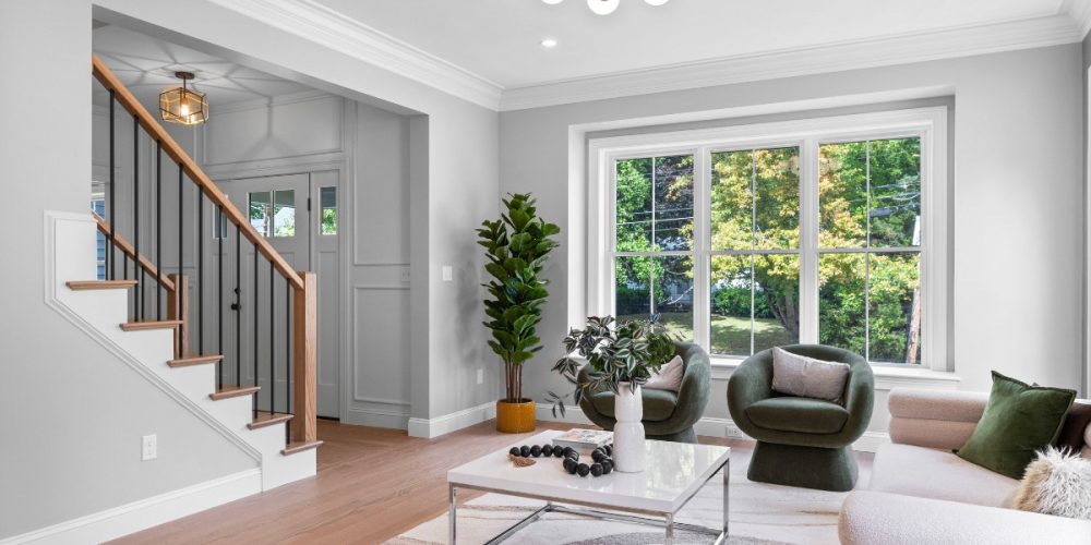 A bright, modern living room designed by Kadilak Homes in Burlington, Massachusetts, featuring Benjamin Moore Collingwood paint on the walls, large windows letting in natural light, green accent chairs, and a sleek staircase with black metal balusters.