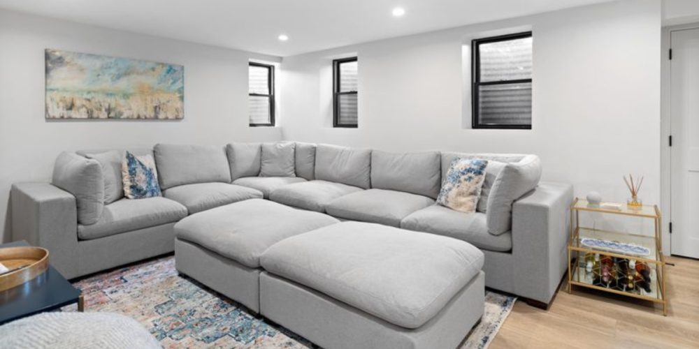 A modern, cozy basement family room featuring a large gray sectional sofa, three small windows allowing natural light, and a stylish gold bar cart. The walls are painted a crisp white, complemented by recessed lighting and a patterned area rug for warmth.