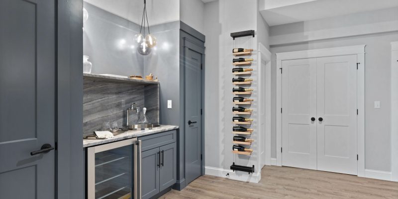 A modern basement wine bar elegantly designed with a sleek gray color scheme. The room features a dark marble countertop with a built-in sink and gray cabinetry, including a wine fridge. Above the countertop, the marble extends to the backsplash, and two stylish pendant lights hang down, providing a warm ambiance. To the left of the image, a full-height wine rack with bottles on display creates a focal point. The background includes double doors with a white finish, enhancing the contemporary look, and the flooring is light wood, contrasting nicely with the darker tones of the furniture.