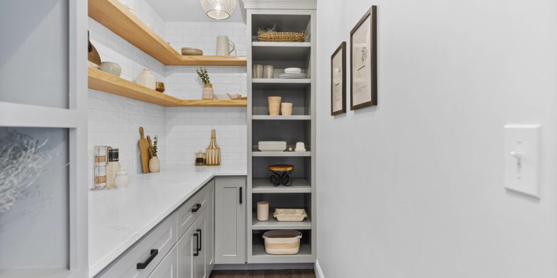 "A modern, well-organized pantry with floating wooden shelves on the left displaying neatly arranged kitchen items, including ceramic jars and a plant. Below, a white countertop runs the length of the space, featuring more kitchen utensils and a stylish gold pendant light above. To the right, a tall gray shelving unit holds a variety of dishes and baskets. The room is finished with a herringbone-patterned dark wood floor, white subway tile backsplash, and framed botanical prints on the wall.