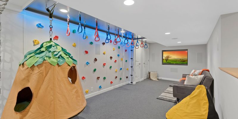 A playful and vibrant children's play area in a basement, featuring a climbing wall with colorful holds and a row of gymnastic rings suspended from the ceiling, set against a backdrop of bright blue lighting. To the left, there's a whimsical tent resembling a tree, perfect for imaginative play. The floor is covered with a grey, soft carpet, providing a safe surface for activities. A comfortable seating area with a dark couch adorned with orange and white pillows sits in the background, facing a large, framed landscape painting of a tree at sunset, adding a touch of serenity to the dynamic space. The room is well-lit with recessed ceiling lights, ensuring a bright and inviting environment.