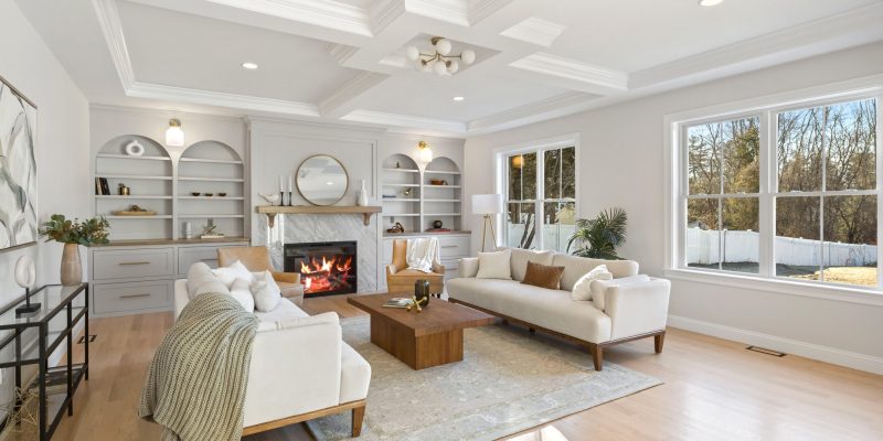 A bright and inviting living room bathed in natural light from large windows, showcasing a crisp white interior with subtle gray accents. Two plush sofas are arranged facing each other, one draped with a cozy knitted blanket, over a light patterned area rug. A solid square coffee table sits between them. An elegant fireplace with a textured surround and white mantelpiece commands attention, adorned with minimal decor and a round mirror above it. White built-in shelving units elegantly display curated decorative items and books. A floor lamp with a white shade provides additional light, complementing the room's serene ambiance. The coffered ceiling with recessed lighting adds depth to the room, enhancing its spacious and luxurious feel. Benjamin Moore Seapearl Oc-19