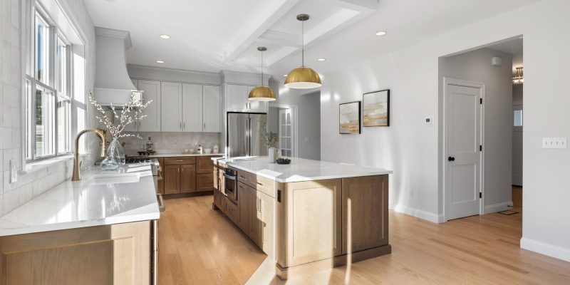 An airy kitchen space with abundant daylight cascading through large windows onto the light hardwood floors. The kitchen boasts a mix of white and natural wood cabinetry, providing a harmonious blend of modern and rustic elements. A central island with a white marble countertop offers additional workspace and storage, complete with a built-in sink and gold faucet. Two large gold pendant lights hang above the island, contributing to the kitchen's elegant aesthetic. White subway tiles create a clean backsplash behind the countertops, complementing the stainless steel appliances, including a prominent double-door refrigerator. Artistic wall pieces adorn the walls, adding a touch of color to the otherwise neutral palette, and the coffered ceiling with recessed lights ensures a well-lit environment. Benjamin Moore cascade mountains 862