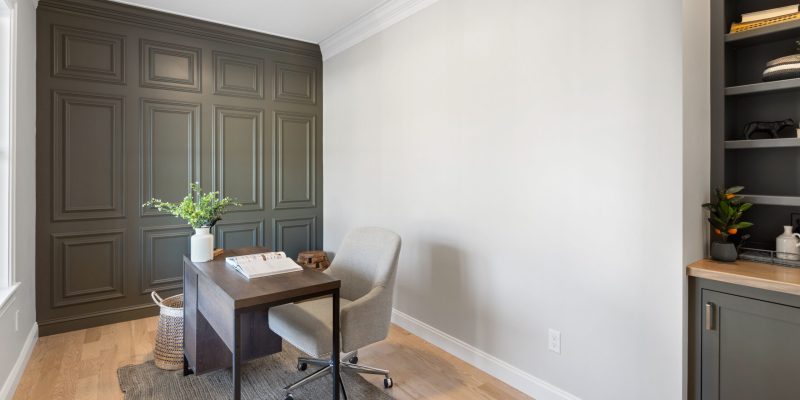 A modern home office with a minimalist design. The room features a dark olive green wainscoting on one wall, creating a sophisticated backdrop. On the right, there's a built-in shelf painted in the same hue, with neatly arranged books and decorative items. The other wall is painted in a light cream color, complementing the dark tones. In the center, a stylish wood and metal desk holds an open book, a white ceramic vase with fresh greenery, and a pair of reading glasses, suggesting a quiet place for reading or work. A comfortable, light gray upholstered chair with a metal base is positioned at the desk, and a woven basket sits nearby on the floor. The natural light from an unseen window softly illuminates the space, highlighting the warm hardwood flooring and the textured gray area rug under the desk area. Benjamin Moore Aegean Olive 1491