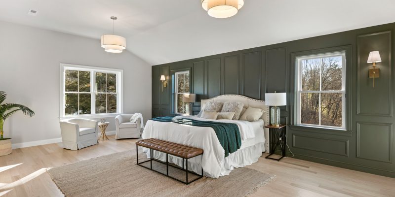A spacious and serene bedroom with a wall of dark green wainscoting providing a dramatic backdrop to an elegant white bed adorned with plush pillows and a green throw. Two large windows allow natural light to stream in, highlighting the light hardwood floors and a large area rug. A tufted leather bench sits at the foot of the bed, and a cozy reading nook is created with two armchairs and a small side table near the window. The room features two stylish drum pendant lights, and on the far side, a thriving potted plant adds a touch of nature to the interior. This bedroom exudes a relaxed, luxurious atmosphere. Ashwood Moss 1484