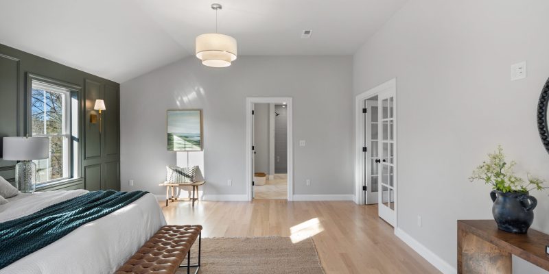 A spacious and airy bedroom featuring a vaulted ceiling with a central drum pendant light. Natural light streams in through windows set in a dark green wainscoted wall. The room is furnished with a large bed covered in white linens and accented with a teal throw blanket, with a tufted leather bench at its foot. To the left, a cozy nook with a wooden chair and a side table offers a place for relaxation. On the right, a dark wood console table hosts a decorative vase with greenery, contributing to the room's natural and calm ambiance. The light hardwood floor is partially covered by a textured area rug, and double doors open to an adjoining room, providing a glimpse into the home's continuity and design. Benjamin Moore Balboa Mist OC-27