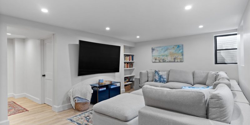 A finished basement living area featuring a large gray sectional sofa centered around a glass coffee table, facing a wall-mounted flat-screen TV. The space is well-lit with recessed lighting and features a small window with blinds on the right, letting in limited natural light. The flooring is light-toned luxury vinyl plank, and the room has a modern, minimalist aesthetic. There is a white built-in bookshelf to the right of the TV filled with books, and a colorful abstract painting hangs on the wall to the right. The room is accented with a traditional patterned area rug and a smaller decorative rug in front of a closed white door on the left. A woven basket with a blanket peeks out from under a small throw blanket on the coffee table, adding a homey touch to the space.