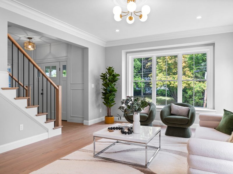 Bright and airy living room with large picture windows, modern white and green furniture, and a cozy seating area. Features an oak and black baluster staircase, white wall paneling, and contemporary gold light fixtures. The open layout offers a seamless flow from the living space to the entryway, creating an inviting and stylish home interior