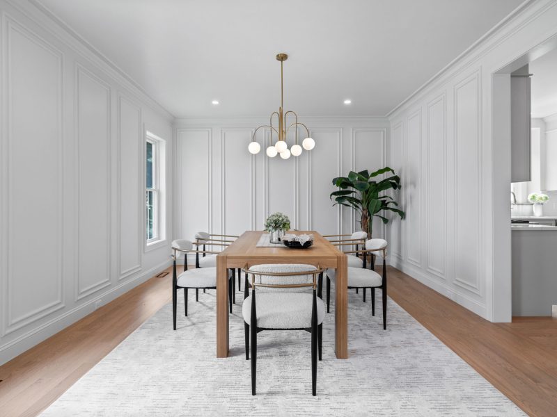 "Elegant dining room featuring a wooden dining table with modern upholstered chairs, white wall paneling, and a brass chandelier. The room is accented by light hardwood floors, large windows providing natural light, and a statement plant in the corner, creating a clean and sophisticated dining space.