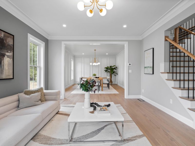 Modern open-concept living and dining room featuring light hardwood floors, a cozy white sofa, and a white coffee table with contemporary decor. The space is accented by black and oak staircase railings, sleek lighting fixtures, and large windows that allow natural light to fill the room. The dining area showcases elegant white wall paneling and a stylish chandelier.