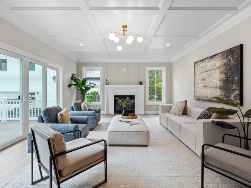 Modern living room featuring an electric fireplace with a white mantel, grasscloth wallpaper, and coffered ceiling. The room includes a beige sectional sofa, blue armchairs, and a large ottoman, with sliding glass doors leading to an outdoor deck. Natural light from large windows and sliding doors brightens the space, while a statement plant adds a touch of greenery.