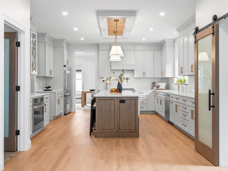 Spacious modern kitchen featuring a large central island with wood cabinetry, light gray shaker cabinets, and brass hardware. The kitchen includes quartz countertops, stainless steel appliances, and a barn-style sliding door leading to the pantry. A unique ceiling design with an oak shiplap insert and pendant lighting adds character to the space.
