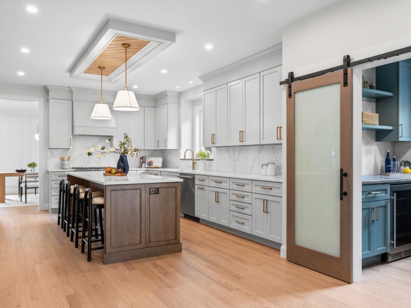 Spacious kitchen featuring Benjamin Moore Pale Oak shaker cabinets, a large wooden island with seating, and brass hardware. The design includes quartz countertops, a marble backsplash, and a barn-style sliding door leading to a pantry with earthy green cabinetry with wood countertops. A unique ceiling detail with oak shiplap and pendant lighting adds warmth to the modern space.