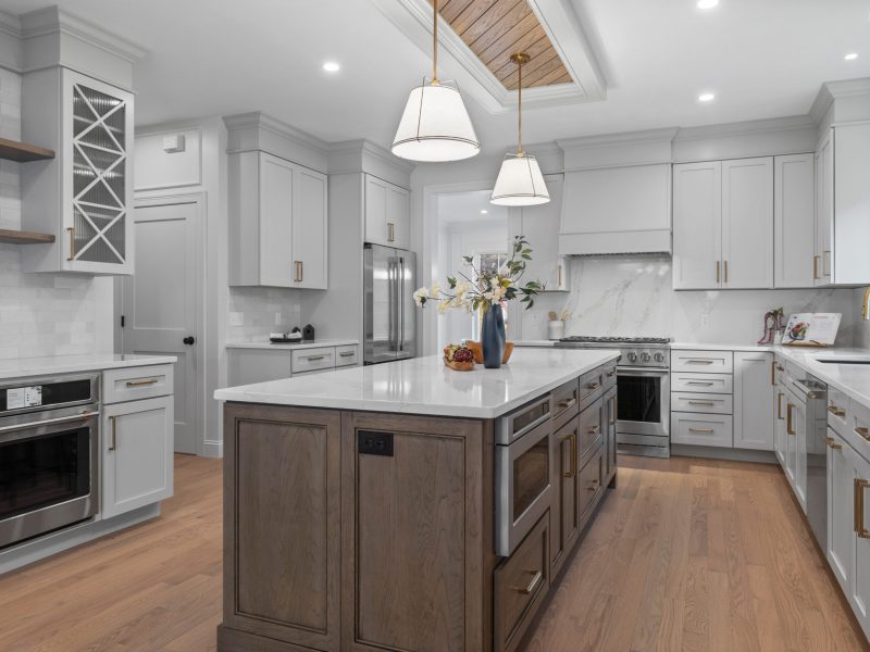 Modern kitchen with a large wooden island, benjamin Moore pale oak shaker cabinets, quartz countertops, and stainless steel appliances. The kitchen features brass hardware, a marble backsplash, and a unique ceiling design with oak shiplap and pendant lighting, creating a stylish and functional space.