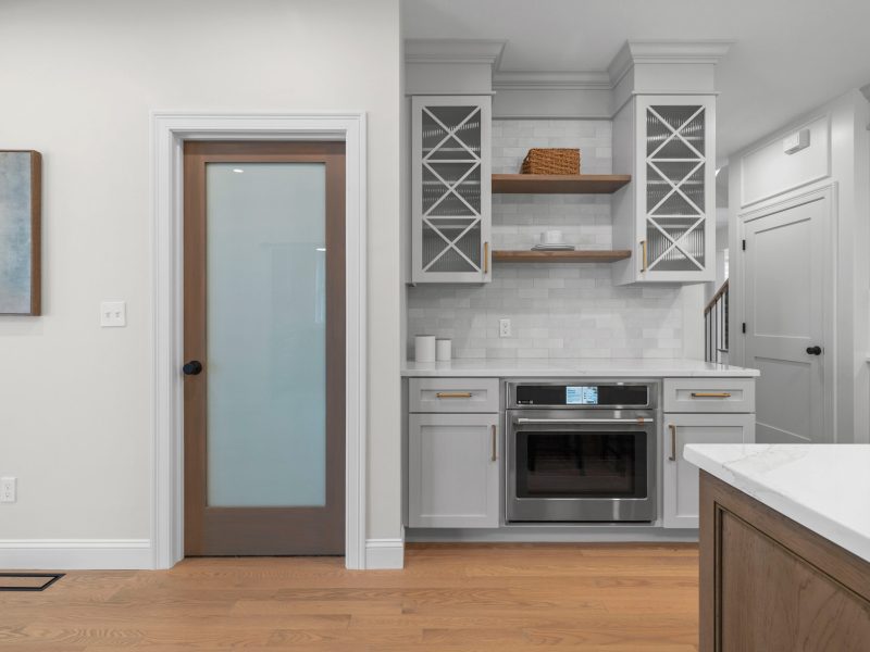 Modern kitchen featuring light gray shaker cabinets with glass fronts, open wooden shelves, and stainless steel appliances. The design includes a frosted glass pantry door and a subway tile backsplash, creating a clean and stylish look.