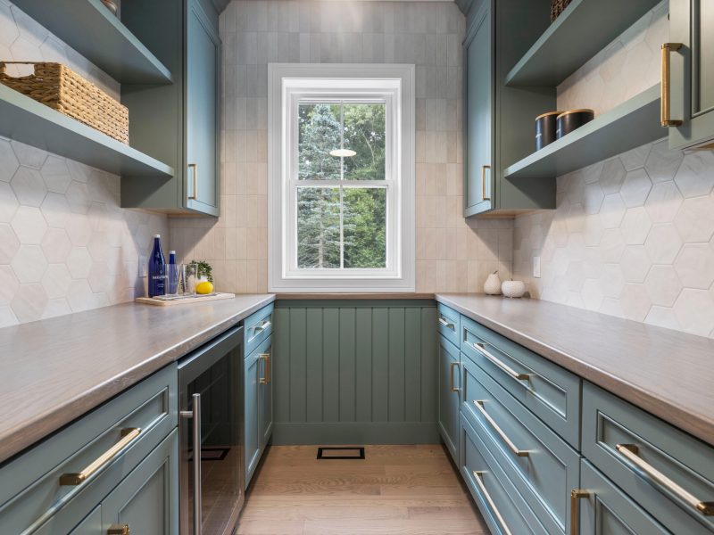Stylish walk-in pantry featuring earthy green shaker cabinets, gold hardware, and a hexagonal tile backsplash. The space includes wooden countertops, open shelving, and a beverage refrigerator beneath the counter. A large window provides natural light, highlighting the elegant design.