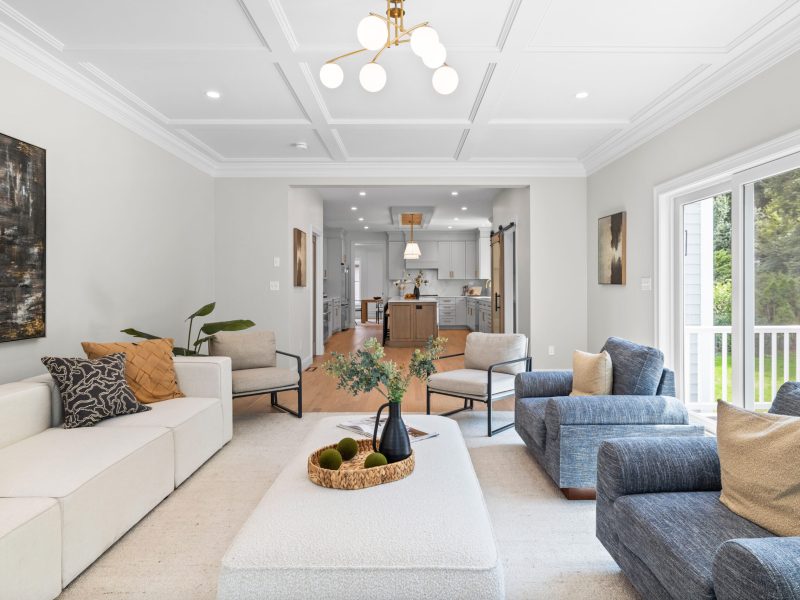 Open-concept living room leading to a modern kitchen, featuring a coffered ceiling, beige sectional sofa, and blue armchairs. The space is filled with natural light from sliding glass doors, offering a seamless indoor-outdoor connection. A large ottoman and contemporary decor add comfort and style, while the view of the kitchen includes a central island and sleek lighting fixtures.