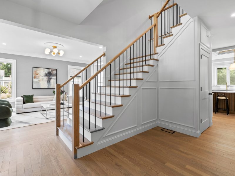 Modern open-concept living room and staircase featuring hardwood floors, black matte balusters, and oak handrails. The clean white panel molding adds elegance to the stairwell, while large windows and contemporary lighting provide natural light and a bright, welcoming atmosphere