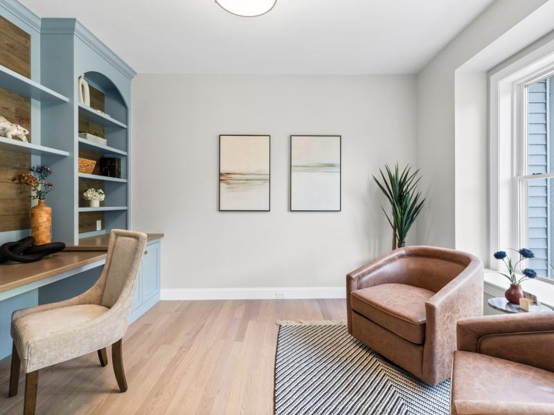 Home office featuring built-in shelves and desk painted in Benjamin Moore Kentucky Haze AC-16. The room includes leather accent chairs, light wood flooring, and large windows that provide natural light. Minimalist decor with neutral tones and wood paneling creates a calm, inviting workspace