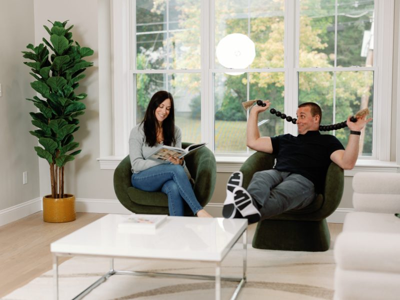 Couple relaxing in a bright living room with large windows, modern green swivel chairs, and a white coffee table. Natural light fills the space, highlighting the cozy seating area and contemporary decor, including a fiddle leaf fig plant in a stylish pot.