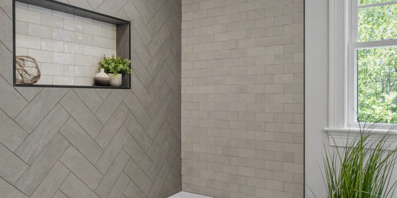 A chic and tranquil bathroom corner is depicted, characterized by its sophisticated tile work and serene ambiance. On the left wall, a striking herringbone pattern of matte grey tiles adds a dynamic and modern touch to the decor. This textured wall contrasts beautifully with the adjacent wall, which features simpler, light grey subway tiles laid in a traditional brick pattern, enhancing the room's depth and dimension. A narrow recessed shelf, framed by a sleek black border, provides a decorative niche, showcasing a tasteful arrangement of bathroom accessories including a spherical nautical-themed decor piece and a couple of vases with lush greenery, adding a touch of organic vibrancy to the space. The corner is further accentuated by the presence of a white bathtub, with its pristine edge visible, inviting relaxation. A large window with white trim on the right side floods the space with natural light and frames a view of verdant trees, reinforcing the bathroom's serene and refreshing atmosphere. The thoughtful design choices suggest a harmonious blend of functionality and style, creating an elegant retreat within the home.